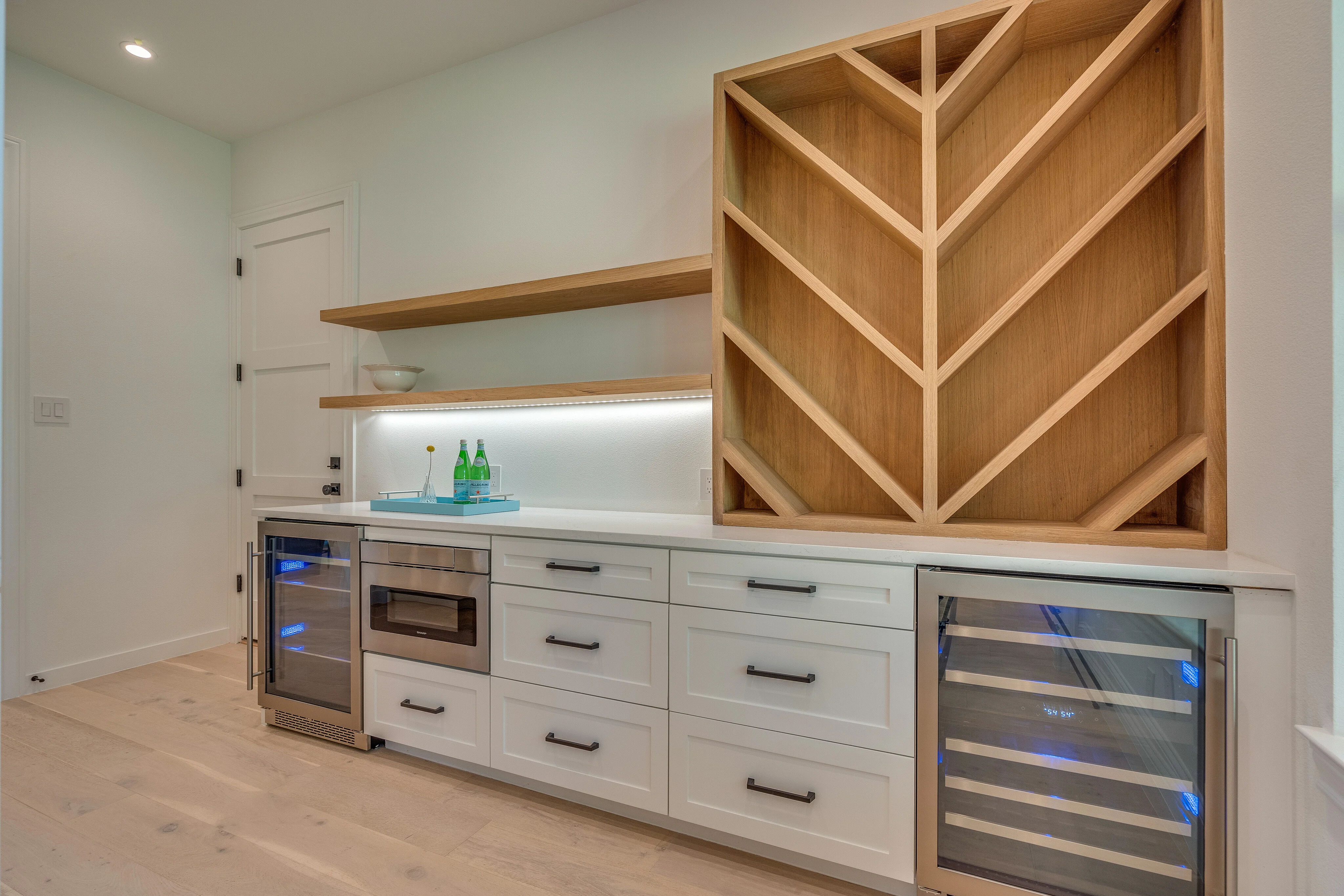Interior beverage station with white cabinetry, pale wood shelving, geometric millwork, and under-shelf lighting