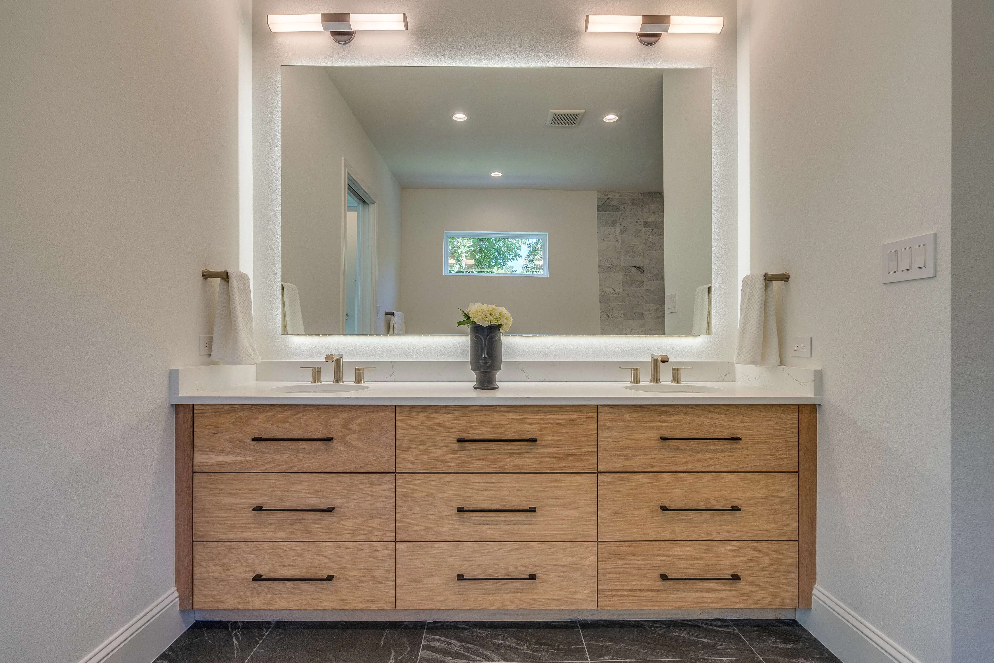 Bathroom with a wide pale-wood vanity, dual sinks, illuminated mirror, and dark stone floor